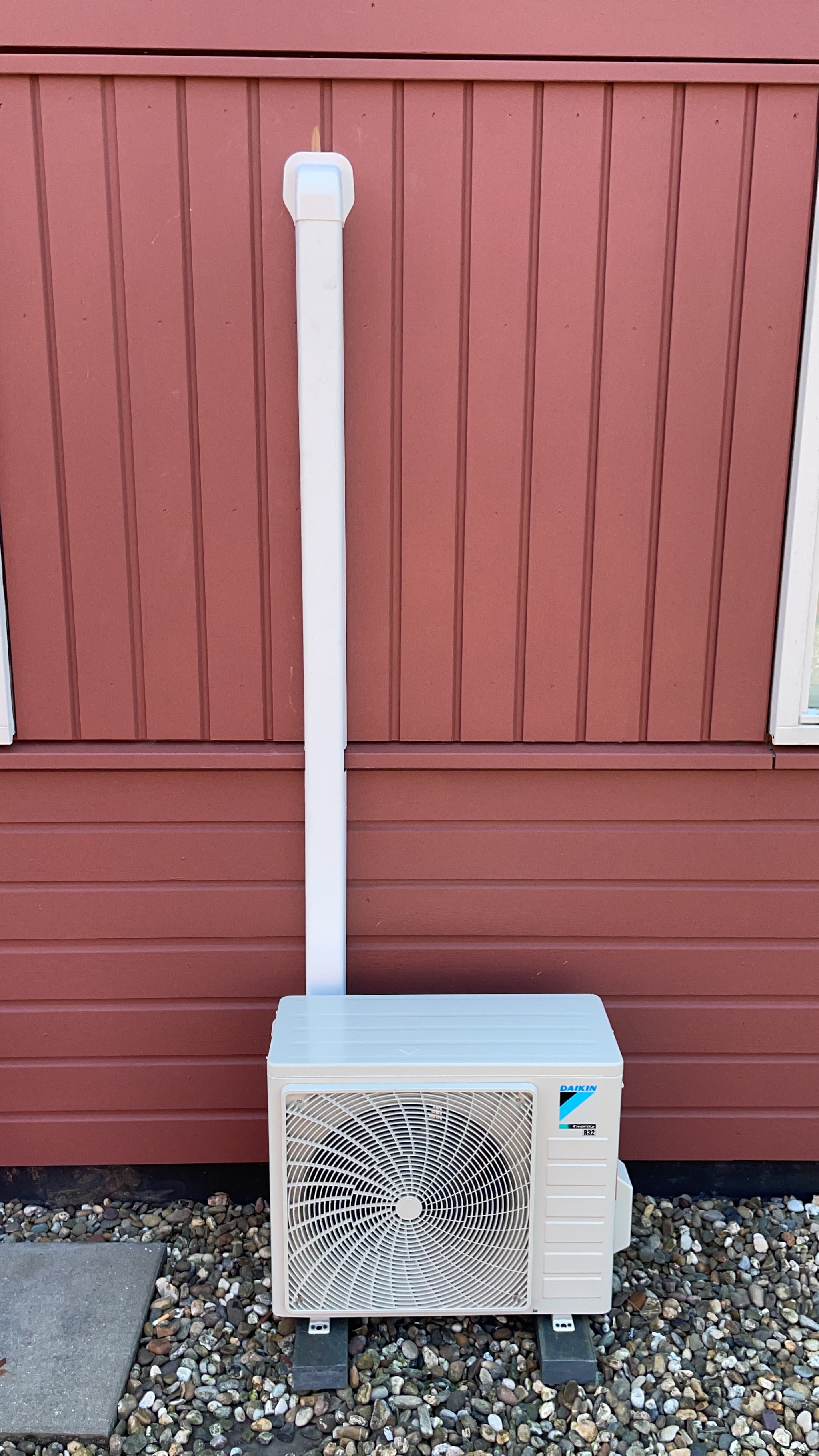 Daikin buitenunit op grind voor een rode houten gevel met witte leidingkoker
